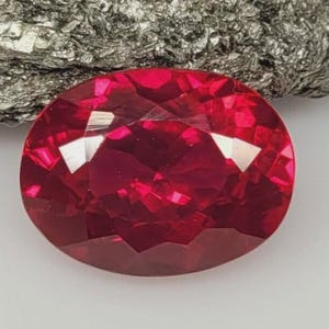 May include: An oval-cut gemstone with a deep, vibrant red colour and multiple facets that reflect light. The stone displays a high level of clarity and brilliance, showcasing a polished surface with sharp geometric edges throughout its structure.