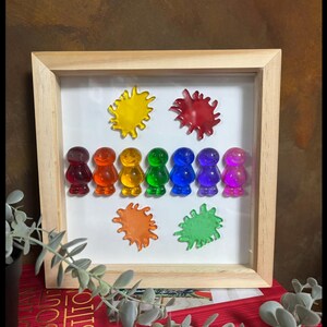 Könnte beinhalten: Ein gerahmtes Kunstwerk mit farbenfrohen, durchscheinenden Figuren und Farbspritzern. Die Figuren sind in einer Reihe angeordnet, jede in einer anderen Farbe des Regenbogens. Die Farbspritzer sind gelb, rot, orange und grün.