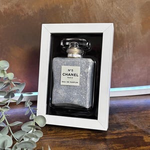 Op de afbeelding: Een rechthoekige parfumfles met een zilveren glitterinterieur, gepresenteerd in een witte lijst. De fles heeft een zwarte dop en een label met de tekst "N°5 CHANEL PARIS EAU DE PARFUM". Op een bruin houten oppervlak.