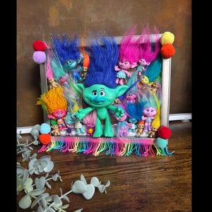 May include: A framed display featuring multiple Troll dolls with colorful hair. The central figure is a large green Troll. The frame is decorated with colorful pom-poms and a rainbow fringe. The background is a mix of other Troll dolls.