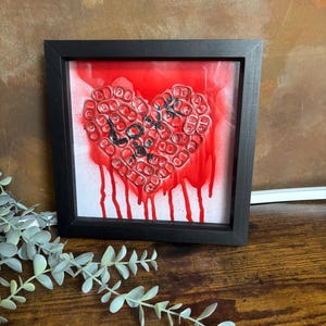 Op de afbeelding: Een ingelijst kunstwerk met een hart van rode blikjes, met het woord "Love" in zwart schrift. De achtergrond is rood met een druipend verfeffect. De zwarte lijst staat op een houten oppervlak.