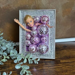 Op de afbeelding: Een ingelijst kunstwerk met een poppenhoofd en armen die uit een cluster van paarse discoballen komen. De zilveren lijst heeft glitteraccenten, tegen een bruine achtergrond. Het kunstwerk heeft een grillig en decoratief thema.