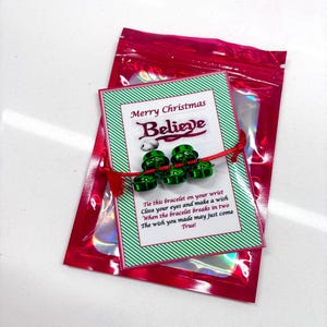 May include: A red string bracelet with three small green jingle bells attached to a rectangular card featuring green and white diagonal stripes. The card text reads Merry Christmas Believe and includes instructions to make a wish.
