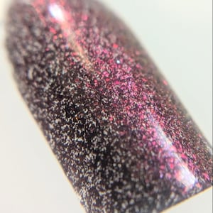 May include: A nail polish swatch featuring a deep black base color with a dense concentration of fine, shimmering particles that shift between dark purple and vibrant magenta tones, creating a textured, multi-dimensional appearance on the surface.