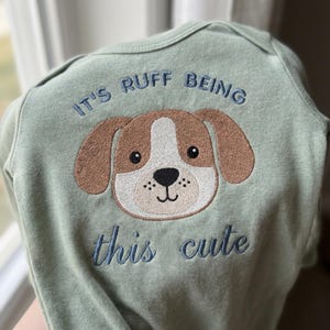 May include: A light green baby onesie with an embroidered dog face. The dog has brown ears, a white face, and black eyes. The text "IT'S RUFF BEING this cute" is embroidered in blue thread.