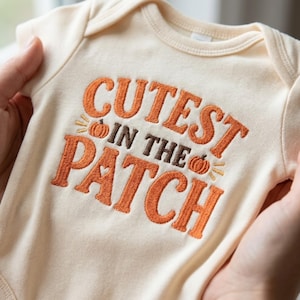 Könnte beinhalten: Ein cremefarbener Babybody mit gesticktem Text CUTEST IN THE PATCH in orangefarbenem und braunem Garn. Das Design enthält zwei kleine orangefarbene Kürbissymbole mit gelben Akzentlinien und eine kleine Herzform innerhalb des Buchstabens A.