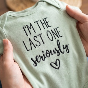 May include: A light green baby onesie with black embroidered text that reads "I'M THE LAST ONE seriously" with a heart symbol. The onesie is held up by a person's hands.