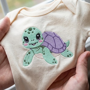 May include: Cream-colored baby onesie featuring an embroidered sea turtle design. The turtle has a green head and limbs, a purple shell, and pink cheeks. The onesie is made of soft fabric, perfect for newborns.