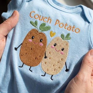 May include: Light blue baby onesie with the embroidered text "Couch Potato" and two smiling potato characters. The potatoes are brown and tan with green sprouts and a yellow heart.
