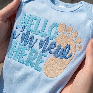 May include: A pale blue baby bodysuit featuring the embroidered phrase "Hello New Here" in blue hues. A tan embroidered footprint is next to the text. The bodysuit is held by a person.