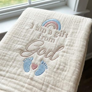 Può includere: Una coperta avvolgente in mussola color crema con il testo ricamato I am a gift from God. Il design include un arcobaleno pastello sopra il testo e due impronte di piedi di neonato azzurre con un piccolo cuore rosa sotto il testo.