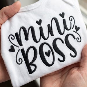 May include: White baby onesie with the words "mini BOSS" embroidered in black script. The design includes heart accents. The onesie is held up by a person's hands.