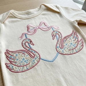 May include: Cream-colored baby onesie featuring an embroidered design of two swans facing each other within a light blue heart. The swans have floral patterned wings and pink outlines. A pink bow is above the heart.