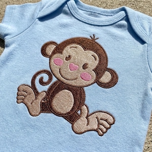 Puede incluir: Un body de bebé azul claro con un diseño de mono bordado. El mono tiene pelaje marrón, mejillas rosas y una nariz en forma de corazón rosa. El body está hecho de tela suave y tiene mangas cortas.