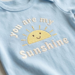 以下が含まれることがあります： 水色のベビー用ロンパース。刺繍された「You are my Sunshine」の文字と、笑顔の黄色い太陽のデザインが特徴です。柔らかい生地で作られており、半袖と丸い襟ぐりです。
