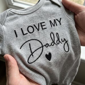 May include: A heather gray infant bodysuit featuring black embroidered text that reads I LOVE MY Daddy with a small black heart icon centered below the word Daddy. The garment has short sleeves and a round neckline with visible stitching.