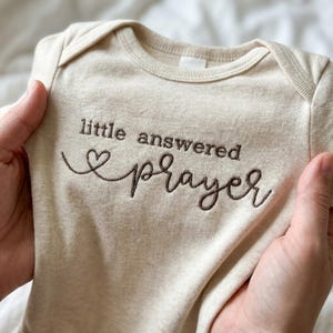 May include: A beige baby onesie with short sleeves. The words "little answered prayer" are embroidered in brown script, with a heart symbol. The onesie is held up by two hands.