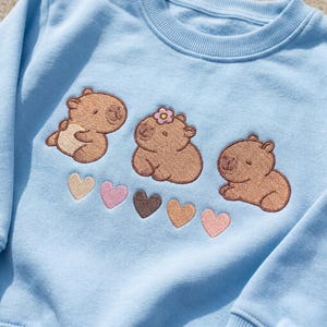 May include: A light blue crewneck sweatshirt featuring an embroidered design of three brown capybaras. The centre capybara wears a small pink flower on its head. Below the capybaras are five small embroidered hearts in shades of beige, pink, and brown.