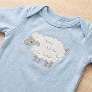 Puede incluir: Un body de bebé azul claro con un diseño de oveja bordada. La oveja tiene un cuerpo blanco texturizado, cabeza gris y patas grises. El body está hecho de material suave y tiene mangas cortas.
