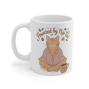 May include: White ceramic mug featuring a cartoon cat in a meditative pose, wearing a pink robe. The mug has the text "Powered by Nip Tea" and a small cup of tea with the text "Catnip Tea".