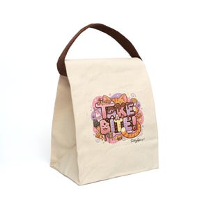 May include: A cream-colored canvas lunch bag with a brown fabric handle. The front features a colorful graphic of an orange cat surrounded by various food items and the text "TAKE A BITE!" in a playful, stylized font with a small signature below.