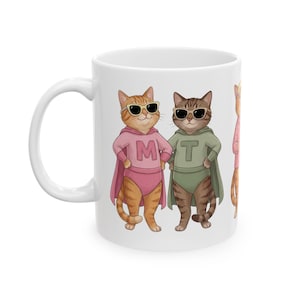 May include: White ceramic mug with a design of cartoon superhero cats. One orange cat wears a pink outfit with the letter "M", the other a brown cat in green with the letter "T". Both cats have sunglasses and capes. The mug has a white handle.