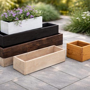 Pode incluir: Várias floreiras de madeira em diferentes tamanhos e cores. A floreira superior é branca e contém flores roxas. Outras são pretas, castanhas e em tons de madeira natural. Uma pequena floreira quadrada também é visível.