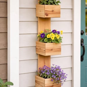 May include: A wooden vertical planter with three rectangular boxes, each filled with colourful flowers. The top box has pink flowers, the middle has yellow and purple, and the bottom has purple flowers. The planter is mounted on a light grey wall.