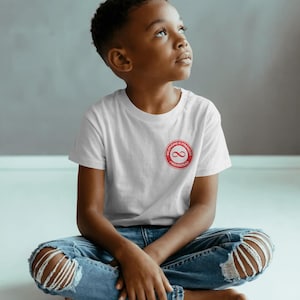 Puede incluir: Una camiseta blanca con un logotipo circular rojo con un símbolo de infinito y las palabras "Belonging is Strength" y "Fredinstead". La camiseta la lleva un niño con vaqueros azules con rodillas desgastadas.