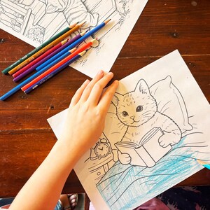 May include: A coloring page featuring a cat reading a book, with a person coloring the image with a blue pencil. Several colored pencils are scattered on the wooden surface. The cat is drawn in a cartoon style.