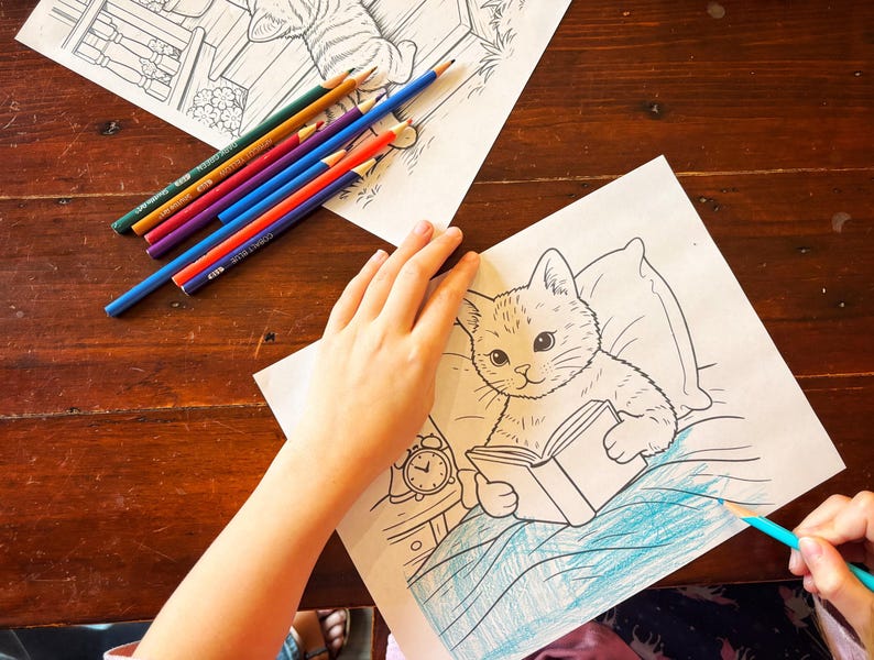 May include: A coloring page featuring a cat reading a book, with a person coloring the image with a blue pencil. Several colored pencils are scattered on the wooden surface. The cat is drawn in a cartoon style.
