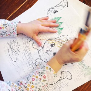 May include: A child's hands coloring a detailed cat illustration with a paintbrush. The coloring book page features a cat in a pond with a lily pad. The child's sleeve has a floral pattern.