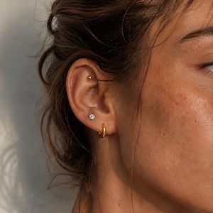 May include: Close-up of an ear with three gold earrings. A small gold ball stud is at the top, a small flower-shaped stud with a clear stone in the middle, and a small gold hoop earring at the bottom.