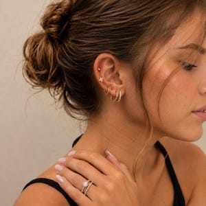 May include: A close-up of a person's ear and hand, displaying gold jewellery. The ear has multiple piercings with small hoop earrings and a flower stud. The hand shows a gold ring with a diamond band and a matching engagement ring.