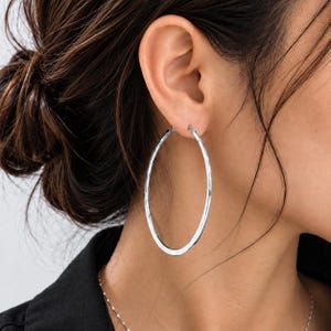 May include: A large, polished silver-tone hoop earring with a smooth, circular design. The earring features a thin, lightweight metal construction that forms a continuous loop, secured through the earlobe with a simple, integrated wire closure.