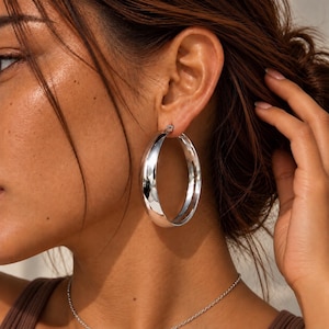 May include: A large, polished silver-tone hoop earring with a thick, rounded profile. The earring features a smooth, reflective metallic surface that catches the light, creating a bright, high-shine appearance on the curved metal structure.