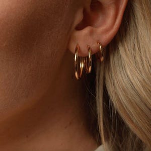 May include: Close-up of a person's ear adorned with three gold hoop earrings of varying sizes. The earrings are shiny and reflect light, creating a warm, golden glow. The background shows blonde hair.