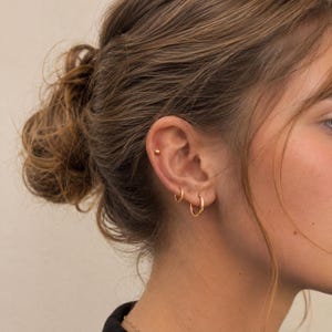 May include: Close-up of a person's ear adorned with gold hoop earrings and a small stud. The earrings are simple, circular designs. The person's hair is pulled back, revealing the ear and jewelry.