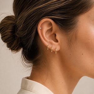 Peut inclure: Un ensemble de trois petites créoles en or poli portées sur le lobe inférieur de l'oreille, accompagné d'une minuscule boucle d'oreille à tige avec une pierre précieuse ronde et transparente située dans le cartilage supérieur de l'oreille.