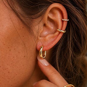 May include: Gold hoop earrings and ear cuffs. The image shows a close-up of an ear adorned with gold jewelry. A pair of small, rectangular hoop earrings are in the earlobe. Two ear cuffs are also visible, one plain and one with small, clear stones.