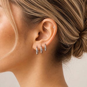 Könnte beinhalten: Drei silberfarbene Creolen in unterschiedlichen Größen am Ohrläppchen, ergänzt durch einen kleinen silberfarbenen Ohrstecker im oberen Knorpelbereich des Ohrs. Der Schmuck zeichnet sich durch eine glatte, polierte und reflektierende metallische Oberfläche aus.