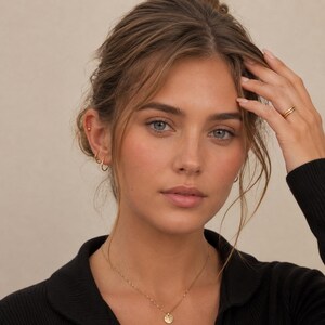May include: A woman wearing gold jewellery, including a delicate necklace with a pendant, small hoop earrings, and a ring. The jewellery complements her complexion and dark top. Her hair is styled up, and she has a natural makeup look.