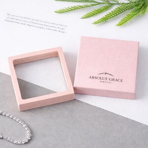 May include: A light pink jewellery box and display case. The box is square with the words "ABSOLUE GRACE JEWELRY" printed on the front. The display case is a square frame with a clear window. A silver bracelet is visible.