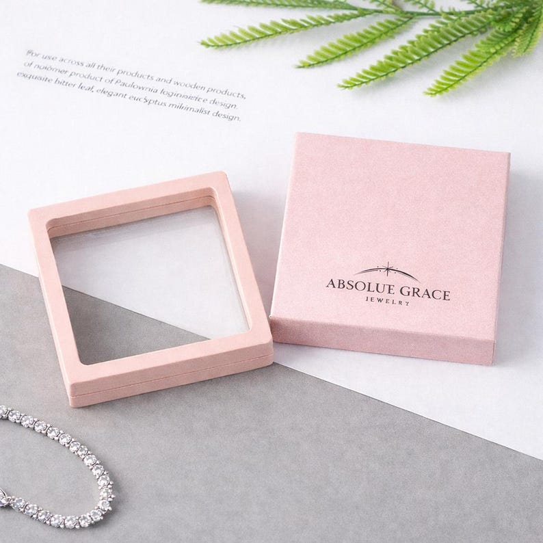 May include: A light pink jewellery box and display case. The box is square with the words "ABSOLUE GRACE JEWELRY" printed on the front. The display case is a square frame with a clear window. A silver bracelet is visible.