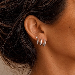 May include: A set of three silver-toned hoop earrings of varying sizes arranged in a row along the earlobe, accompanied by a single small stud earring positioned above the hoops. The jewelry features a smooth, polished metallic finish.