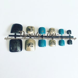 Puede incluir: Un conjunto de diez envolturas para uñas de los pies con temática egipcia. Las envolturas presentan diseños en negro y dorado del Ojo de Horus, pirámides y escarabajos. El texto "Dynasty - Toes - @fantasianailsandmakeup" es visible en la imagen.
