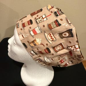 May include: A beige scrub cap featuring a repeating pattern of coffee cups, coffee beans, and coffee drinks. The coffee cups are in various styles, including to-go cups, mugs, and cups with saucers. The cap is on a white mannequin head.