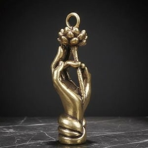 Puede incluir: Una escultura de metal dorado de una mano sosteniendo una flor de loto. La mano está intrincadamente detallada, con los dedos delicadamente colocados alrededor del tallo de la flor. La escultura tiene un lazo en la parte superior para colgar.
