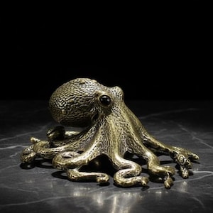 Könnte beinhalten: Eine bronzefarbene Oktopus-Figur mit strukturierten Details. Die Skulptur hat einen abgerundeten Kopf und acht gekräuselte Tentakel. Der Oktopus ist vor einem dunklen Hintergrund platziert, der seine metallische Oberfläche und sein aufwendiges Design hervorhebt.