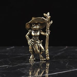 May include: A bronze-colored figurine of a frog-like character wearing a wide-brimmed hat and holding a staff. The figure is detailed with intricate clothing and facial features, standing on a dark surface.